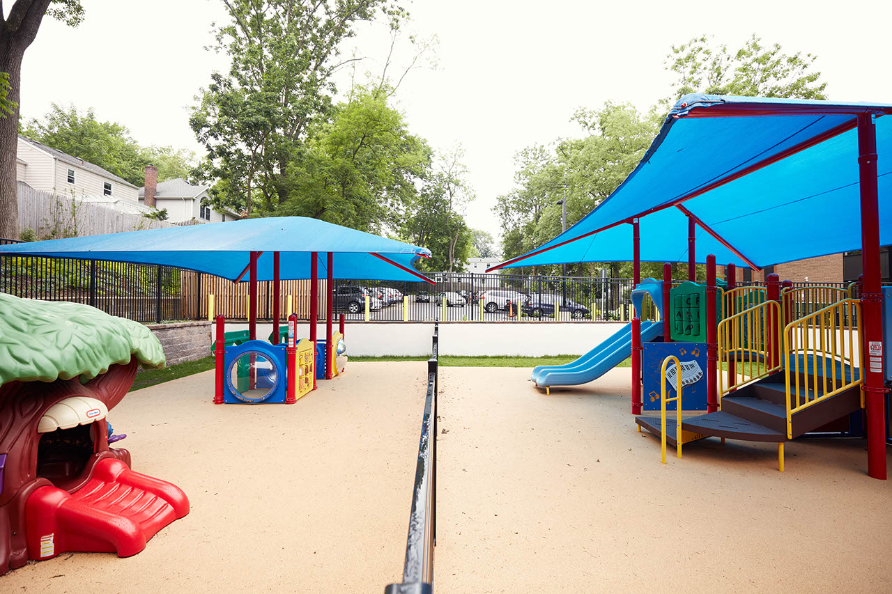 A large, outdoor play space with infant and toddler playgrounds on one side of a fence and a preschool/school-age playground on the other.